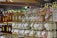 Glass jars filled with colorful substances are neatly arranged on wooden shelves along with bottles of honey. Labels with tags are attached to the jars and bottles, suggesting they are homemade or artisanal products. The jars contain pastel-colored substances, possibly candles or preserved items. The setting appears to be a market or a store.