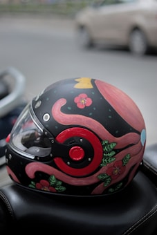A motorcycle helmet with a striking design featuring pink tentacles, green leaves, and red flowers set against a black background. The helmet is placed on a black surface and there is a blurred beige car in the background.