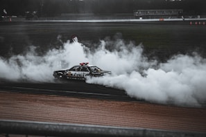 A police car performing a high-speed maneuver on a race track, generating a large amount of white smoke. The background shows a blurred audience and racetrack facilities. The vehicle is decorated with various decals and is fully engaged in a drift.