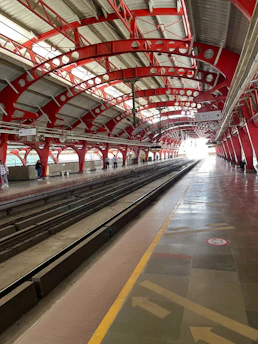 A spacious train station platform with an overhead steel structure featuring red arches and beams. The space is well-lit by natural light coming from large windows and the open end of the platform. The floor is a polished surface with directional arrows and safety markings. A few people are scattered along the platform, waiting.
