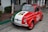 A small vintage car is parked on the street, featuring a tricolor paint job with green, white, and red, resembling the Italian flag. The hood displays an image, and the side is marked with a logo that includes the word 'AutoVino'.