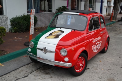 A small vintage car is parked on the street, featuring a tricolor paint job with green, white, and red, resembling the Italian flag. The hood displays an image, and the side is marked with a logo that includes the word 'AutoVino'.