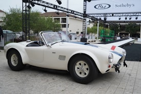 A classic white sports car is parked outdoors on a paved area, framed by green trees and modern architectural structures in the background. A ramp and stage setup, along with a banner reading 'Sotheby's,' suggests an auction or event setting. The vehicle has blue racing stripes and chrome details.