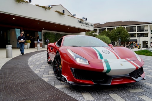 A sleek red sports car with white and green stripes is parked in front of a hotel entrance. The car is positioned on a paved driveway, displaying its front details prominently. Several people are visible in the background near the hotel entrance, with modern architecture and potted plants enhancing the scene.