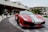 A sleek red sports car with white and green stripes is parked in front of a hotel entrance. The car is positioned on a paved driveway, displaying its front details prominently. Several people are visible in the background near the hotel entrance, with modern architecture and potted plants enhancing the scene.