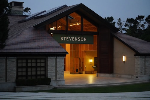 An architectural structure with a wooden roof and illuminated entrance, labeled with the word 'Stevenson'. The building has a modern design with clean lines and large windows. The surroundings include a few trees, contributing to a serene and inviting atmosphere.