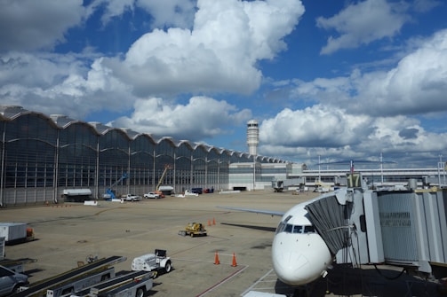 A large airport terminal with a modern architectural design featuring a wavy roofline and expansive glass windows. The runway area is visible with a parked airplane connected to a jet bridge. Various ground vehicles and equipment, such as baggage carts and cargo loaders, are scattered around. The sky is bright with large, fluffy clouds.
