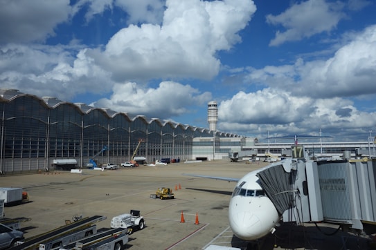 A large airport terminal with a modern architectural design featuring a wavy roofline and expansive glass windows. The runway area is visible with a parked airplane connected to a jet bridge. Various ground vehicles and equipment, such as baggage carts and cargo loaders, are scattered around. The sky is bright with large, fluffy clouds.