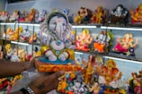 A hand holds a colorful statue of the Hindu deity Ganesha, adorned with intricate jewels and vibrant clothing. The background is filled with various other Ganesha idols displayed on shelves, each embellished with distinct colors and decorations.