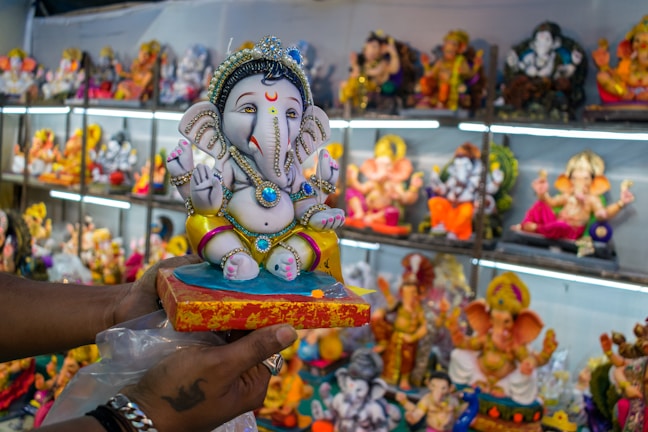 A hand holds a colorful statue of the Hindu deity Ganesha, adorned with intricate jewels and vibrant clothing. The background is filled with various other Ganesha idols displayed on shelves, each embellished with distinct colors and decorations.
