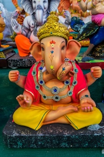 A colorful statue of, featuring intricate details and vibrant colors. It is adorned with jewelry, and the figure is sitting cross-legged with its hands positioned in symbolic gestures. The backdrop includes other similarly styled figures, suggesting a display of religious or cultural significance.