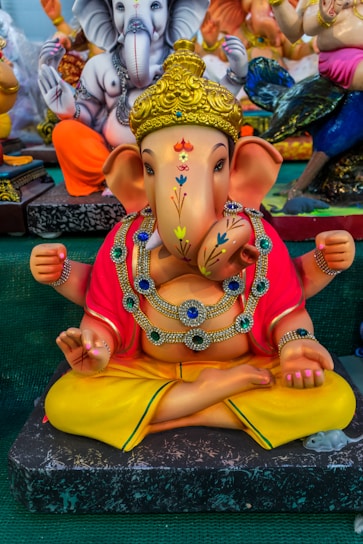 A colorful statue of, featuring intricate details and vibrant colors. It is adorned with jewelry, and the figure is sitting cross-legged with its hands positioned in symbolic gestures. The backdrop includes other similarly styled figures, suggesting a display of religious or cultural significance.