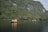 Several small boats with people wearing life vests are floating on a calm, wide river. The backdrop is a lush, green mountain with dense foliage and rugged cliffs. The sky is clear, providing a serene and picturesque landscape.