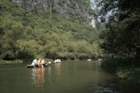 People wearing life jackets are paddling small boats on a calm river surrounded by lush greenery and tall cliffs. The scene conveys a sense of peace and adventure as they explore the serene natural environment.