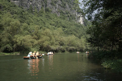 People wearing life jackets are paddling small boats on a calm river surrounded by lush greenery and tall cliffs. The scene conveys a sense of peace and adventure as they explore the serene natural environment.