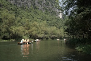 People wearing life jackets are paddling small boats on a calm river surrounded by lush greenery and tall cliffs. The scene conveys a sense of peace and adventure as they explore the serene natural environment.