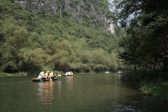 People wearing life jackets are paddling small boats on a calm river surrounded by lush greenery and tall cliffs. The scene conveys a sense of peace and adventure as they explore the serene natural environment.