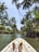 A person is kayaking through a narrow waterway surrounded by lush green palm trees. The view is from the inside of the kayak, with the paddler’s feet visible. The sky is clear, and the sun is shining brightly, creating a calm tropical atmosphere.