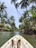 A person is kayaking through a narrow waterway surrounded by lush green palm trees. The view is from the inside of the kayak, with the paddler’s feet visible. The sky is clear, and the sun is shining brightly, creating a calm tropical atmosphere.