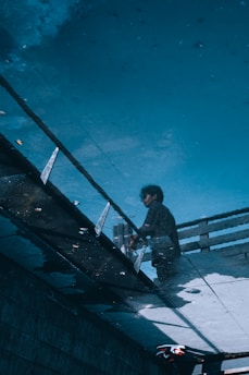 A reflection of a person standing next to a railing, captured in a puddle on the pavement. The image showcases an unusual perspective, blending the reflection with the surrounding urban environment. The captured scene includes geometric shapes from the pavement and railings, creating a visually intriguing composition.