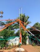 A flagpole stands in the center with the Indian national flag flowing. Strings of alternating orange, white, and green colors are attached to the pole, extending outward like rays. The background includes potted plants, a small building, a parked scooter, and lush green trees against a clear blue sky.