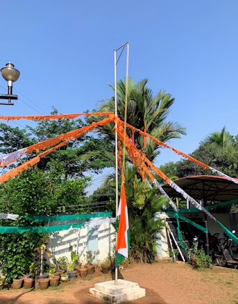 A flagpole stands in the center with the Indian national flag flowing. Strings of alternating orange, white, and green colors are attached to the pole, extending outward like rays. The background includes potted plants, a small building, a parked scooter, and lush green trees against a clear blue sky.