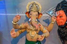 A statue of Ganesha, the elephant-headed Hindu deity, adorned with intricate gold jewelry and a green cloth around the waist. Ganesha is depicted with multiple arms, holding various objects, and features a serene expression. Beside Ganesha, there is a depiction of another deity with a red face and intense expression, holding a silver item.