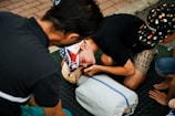 A group of individuals, including a child wearing a cap with a colorful design, are practicing CPR on a dummy. The scene appears to take place outdoors on a textured black mat. The participants are focused on their task, with one child leaning in, presumably to perform mouth-to-mouth resuscitation.