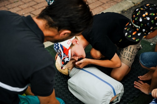 A group of individuals, including a child wearing a cap with a colorful design, are practicing CPR on a dummy. The scene appears to take place outdoors on a textured black mat. The participants are focused on their task, with one child leaning in, presumably to perform mouth-to-mouth resuscitation.