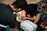 A group of individuals, including a child wearing a cap with a colorful design, are practicing CPR on a dummy. The scene appears to take place outdoors on a textured black mat. The participants are focused on their task, with one child leaning in, presumably to perform mouth-to-mouth resuscitation.