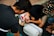 A group of individuals, including a child wearing a cap with a colorful design, are practicing CPR on a dummy. The scene appears to take place outdoors on a textured black mat. The participants are focused on their task, with one child leaning in, presumably to perform mouth-to-mouth resuscitation.