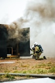 Two firefighters, wearing helmets and protective gear, are kneeling on the ground near a dark structure emitting thick smoke. A hose is lying on the grass nearby, and the scene appears to be part of a firefighting training exercise.