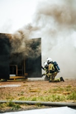 Two firefighters, wearing helmets and protective gear, are kneeling on the ground near a dark structure emitting thick smoke. A hose is lying on the grass nearby, and the scene appears to be part of a firefighting training exercise.