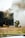 Two firefighters, wearing helmets and protective gear, are kneeling on the ground near a dark structure emitting thick smoke. A hose is lying on the grass nearby, and the scene appears to be part of a firefighting training exercise.