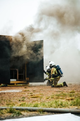 Two firefighters, wearing helmets and protective gear, are kneeling on the ground near a dark structure emitting thick smoke. A hose is lying on the grass nearby, and the scene appears to be part of a firefighting training exercise.