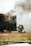 Two firefighters, wearing helmets and protective gear, are kneeling on the ground near a dark structure emitting thick smoke. A hose is lying on the grass nearby, and the scene appears to be part of a firefighting training exercise.