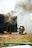Two firefighters, wearing helmets and protective gear, are kneeling on the ground near a dark structure emitting thick smoke. A hose is lying on the grass nearby, and the scene appears to be part of a firefighting training exercise.
