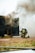 Two firefighters, wearing helmets and protective gear, are kneeling on the ground near a dark structure emitting thick smoke. A hose is lying on the grass nearby, and the scene appears to be part of a firefighting training exercise.