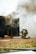 Two firefighters, wearing helmets and protective gear, are kneeling on the ground near a dark structure emitting thick smoke. A hose is lying on the grass nearby, and the scene appears to be part of a firefighting training exercise.