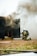 Two firefighters, wearing helmets and protective gear, are kneeling on the ground near a dark structure emitting thick smoke. A hose is lying on the grass nearby, and the scene appears to be part of a firefighting training exercise.