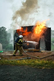 A firefighter in protective gear is actively extinguishing a fire using a hose, with flames and smoke emanating from a burning structure.