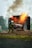 A firefighter in protective gear is actively extinguishing a fire using a hose, with flames and smoke emanating from a burning structure.
