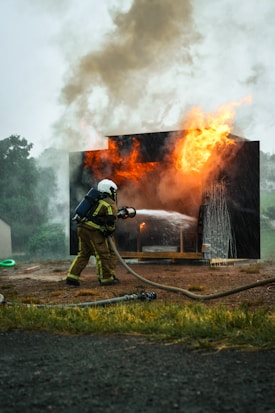 A firefighter in protective gear is actively extinguishing a fire using a hose, with flames and smoke emanating from a burning structure.