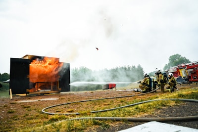 Firefighters in yellow protective suits spray water towards a burning black structure. Flames are visible rising from the building, with smoke billowing into the sky. Fire hoses are laid out on the ground, and a red fire truck is parked in the background. The setting appears to be an outdoor area surrounded by trees.