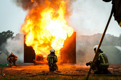 Firefighters in protective gear are actively responding to a large, intense fire. Thick smoke and bright flames create a dramatic scene. The environment appears to be outdoors, with mist or rain adding to the intensity.