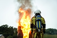 A firefighter wearing protective gear stands facing a large, intense fire with flames and smoke billowing upwards. The firefighter's back is towards the camera, and they are equipped with an oxygen tank. The surrounding area includes trees and a small building in the background.