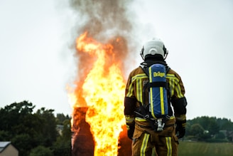 A firefighter wearing protective gear stands facing a large, intense fire with flames and smoke billowing upwards. The firefighter's back is towards the camera, and they are equipped with an oxygen tank. The surrounding area includes trees and a small building in the background.