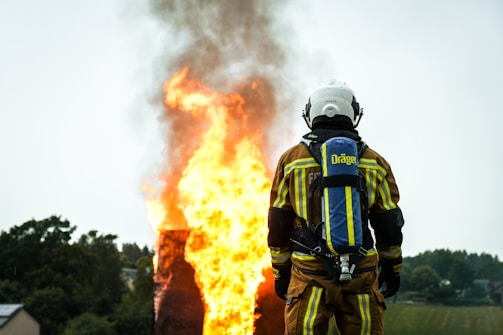A firefighter wearing protective gear stands facing a large, intense fire with flames and smoke billowing upwards. The firefighter's back is towards the camera, and they are equipped with an oxygen tank. The surrounding area includes trees and a small building in the background.