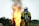 A firefighter wearing protective gear stands facing a large, intense fire with flames and smoke billowing upwards. The firefighter's back is towards the camera, and they are equipped with an oxygen tank. The surrounding area includes trees and a small building in the background.