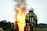 A firefighter wearing protective gear stands facing a large, intense fire with flames and smoke billowing upwards. The firefighter's back is towards the camera, and they are equipped with an oxygen tank. The surrounding area includes trees and a small building in the background.
